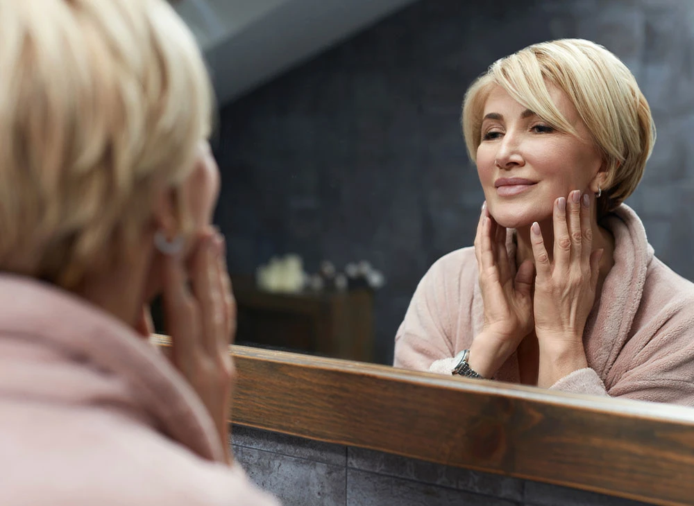 middle aged woman looking in mirror smiling | Ottawa Plastic Surgery in Ottawa, Canada